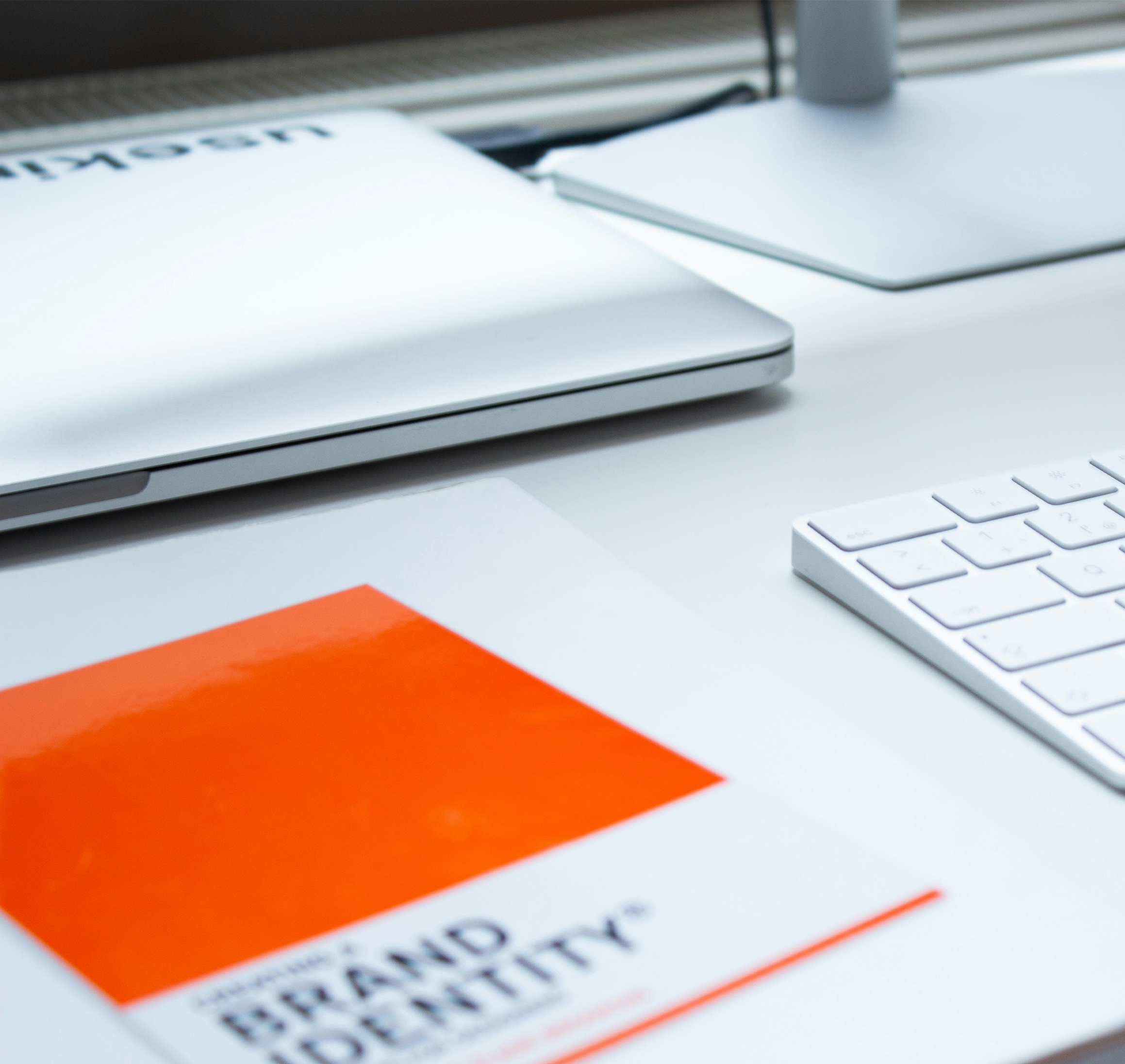 white desktop with an Apple keyboard, a laptop and monitor. Has an orange booklet with the words brand Identity on it.